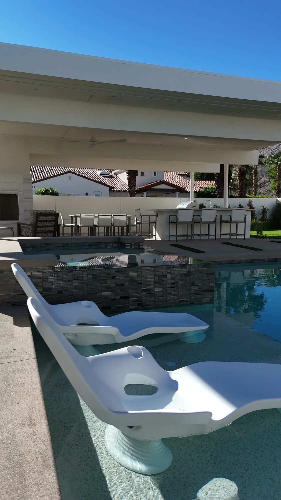 White in-water loungers on a sun shelf beside a raised spa and covered outdoor kitchen